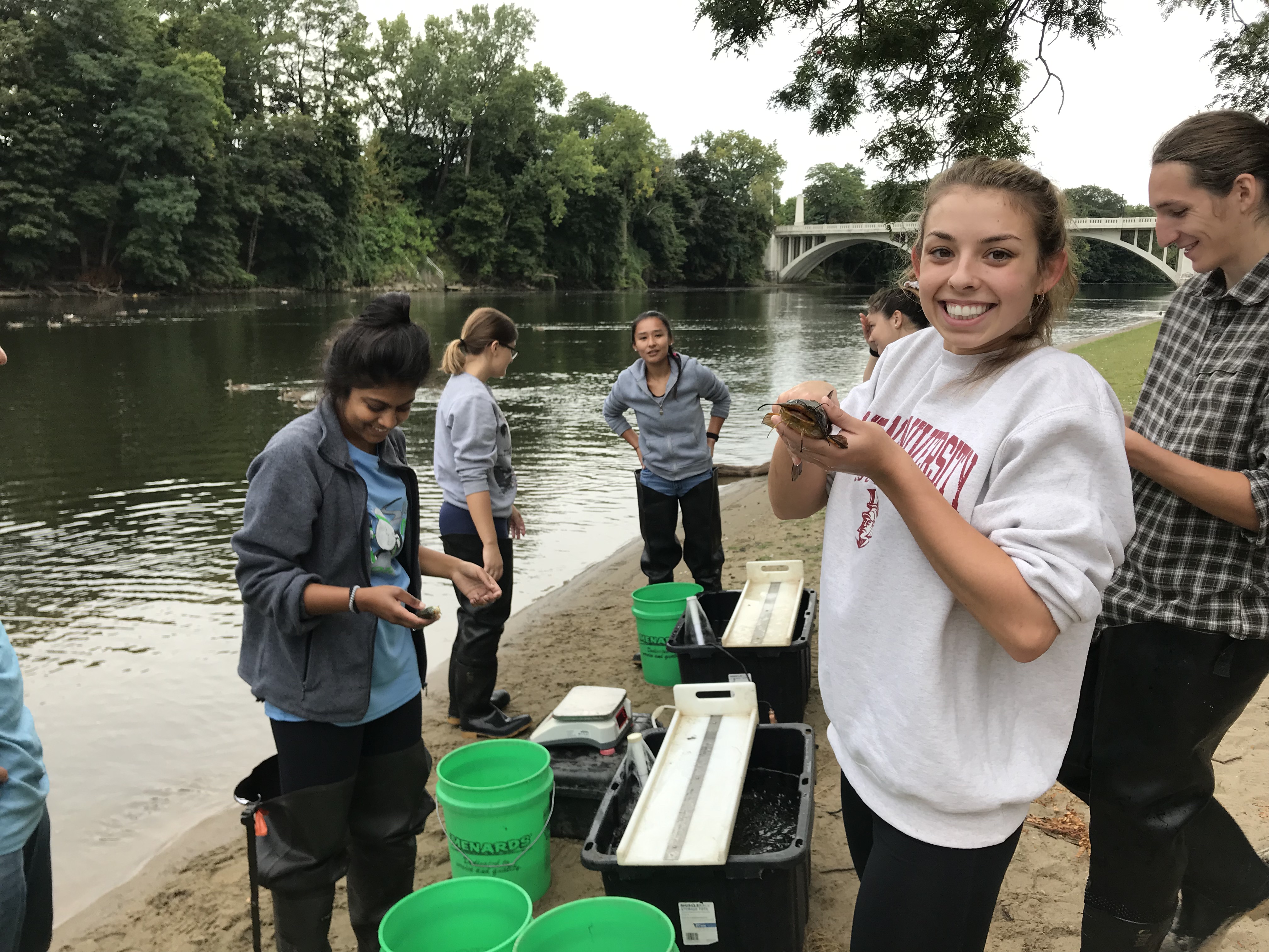 Girl holding catfish and smiling on riverbank with other Field and Laboratory Ecology students